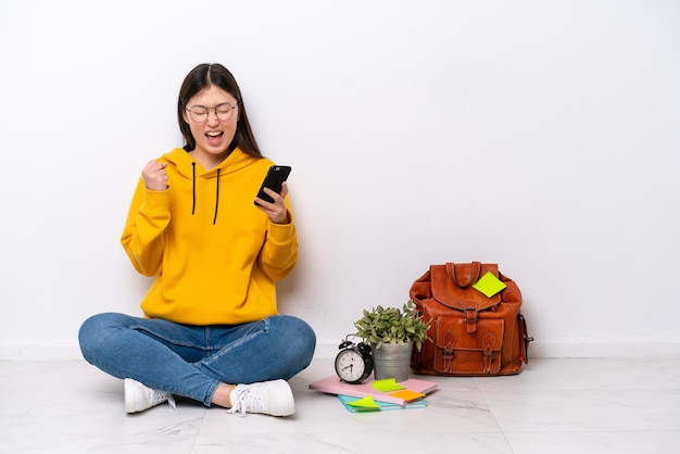 Młoda chińska studentka siedząca na podłodze na białej ścianie z telefonem w pozycji zwycięstwa