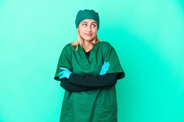 Młoda chirurg urugwajska kobieta w zielonym mundurze na białym tle na niebieskim tle robi gest wątpliwości podczas podnoszenia ramion