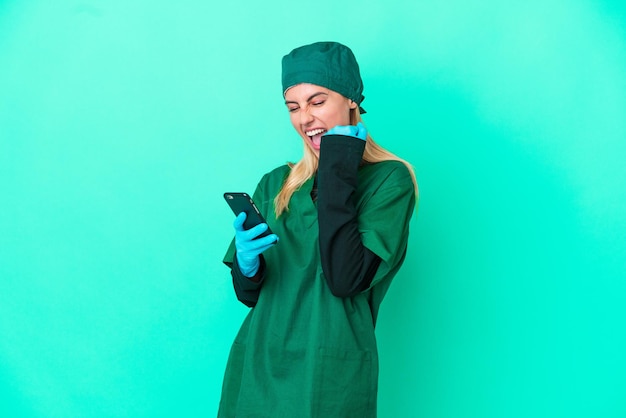 Młoda chirurg urugwajska kobieta w zielonym mundurze na białym tle na niebieskim tle z telefonem w pozycji zwycięstwa