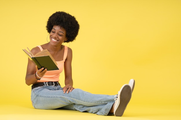 Młoda czarna dama z fryzurą afro siedząca na podłodze szczęśliwa i śmiejąca się podczas czytania książki