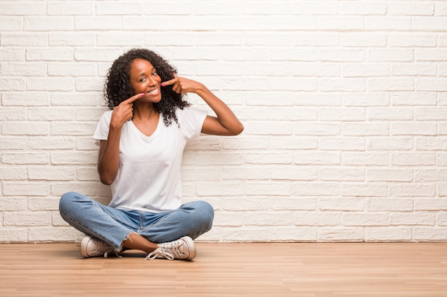 Młoda czarna kobieta siedzi na drewnianej podłodze uśmiecha się, wskazując usta, koncepcja idealnych zębów, białe zęby, ma pogodną i jowialną postawę