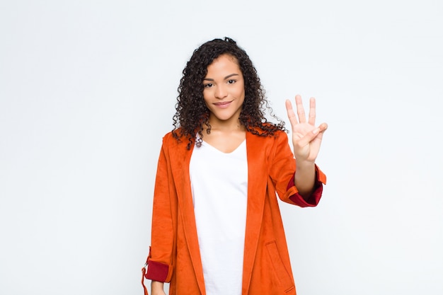 Młoda czarna kobieta uśmiechnięta i wyglądająca przyjaźnie, pokazująca numer trzy lub trzeci z ręką do przodu, odliczający na białej ścianie