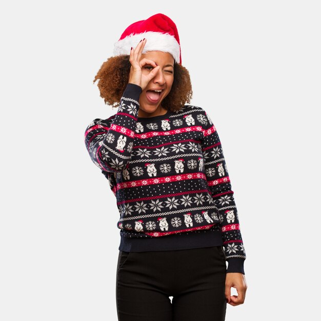 Młoda czarna kobieta w modny sweter christmas z wydruku pewnie robi ok gest na oko