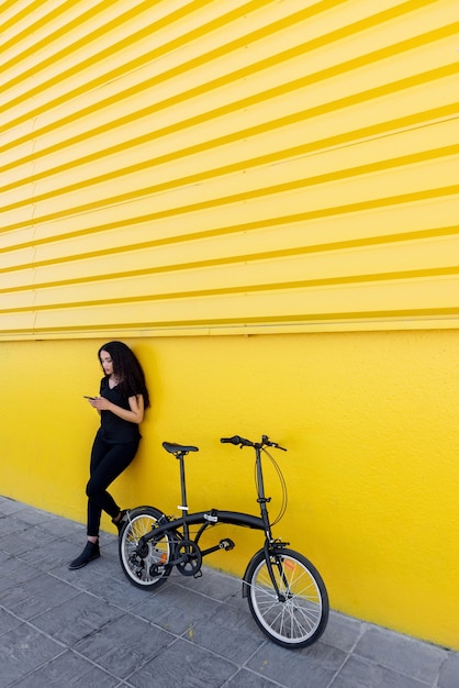 Młoda czarna kobieta z długimi włosami i czarnym strojem z telefonem stojącym obok roweru przed żółtą ścianą