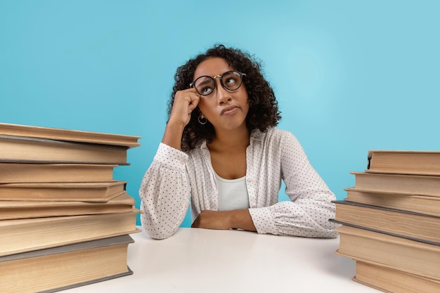 Młoda czarna studentka w okularach siedząca przy biurku ze stosami książek marząca o znudzeniu