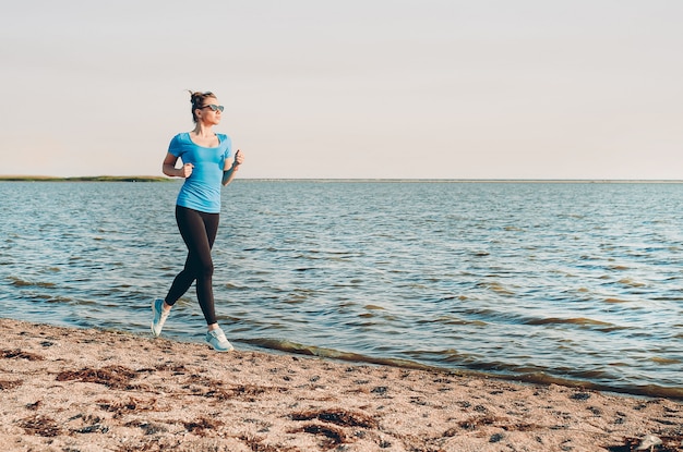 Młoda dama działa. Kobieta lekkoatletka uruchomiony w słoneczny letni piasek na plaży. Trening w pobliżu oceanu wybrzeża morskiego. Piękna, dopasowana dziewczyna. Fitness model kaukaski pochodzenie etniczne na zewnątrz. Ćwiczenia odchudzające. Jogging.