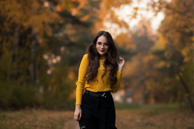Młoda długowłosa brunetka w żółtym swetrze z dzianiny i okularach, patrząc na kamerę, stojąc przed niewyraźnymi liśćmi jesiennych drzew w parku