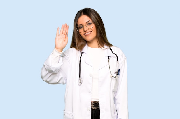 Młoda doktorska kobieta salutuje z ręką z szczęśliwym wyrażeniem na odosobnionym błękitnym tle