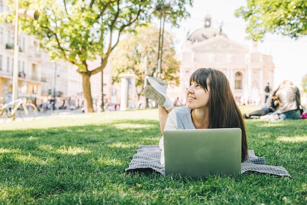 Młoda dorosła kobieta kłaść z laptopem w parku miejskim na zielonej trawie