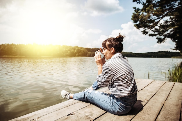 Młoda dorosła kobieta siedząca na drewnianym doku pije kawę i patrzy na jezioro