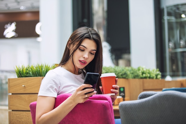 Młoda dorosła piękna brunetka dziewczyna szczęśliwy siedząc w kawiarni z kawą i telefonem komórkowym