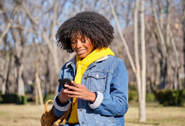 Młoda dziewczyna afro uśmiecha się patrząc na telefon komórkowy w parku