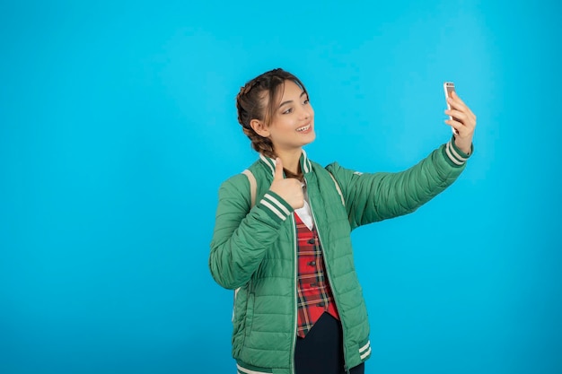 Młoda dziewczyna kolegium biorąc selfie na niebieskim tle. Zdjęcie wysokiej jakości