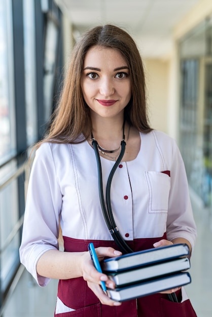 Młoda dziewczyna lekarz w białym fartuchu medycznym z niebieską książką w dłoniach na szpitalnym korytarzu