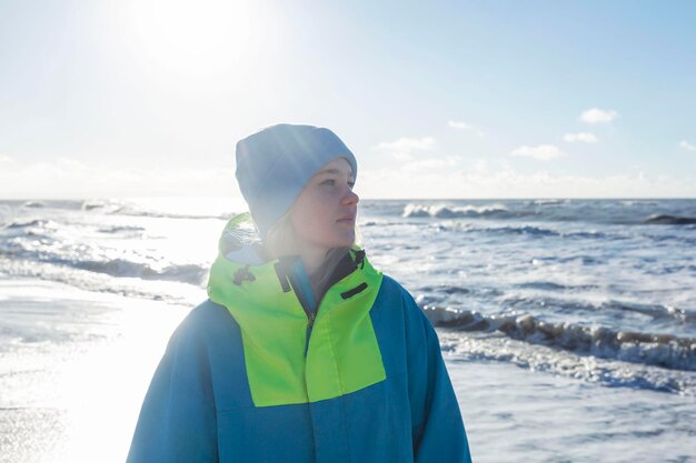 Młoda dziewczyna na morzu w słoneczny zimowy dzień Piękna blondynka w niebieskim kapeluszu i jasnej kurtce sportowej Aktywna rekreacja i spacery