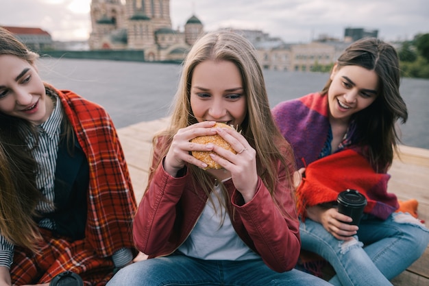 Młoda dziewczyna o gorących oczach zjada hamburgera. Spotkanie ze znajomymi z fast foodami na wynos. Przejadanie się i niewydolność diety, wahania nastroju w jedzeniu. Zamknij zdjęcie