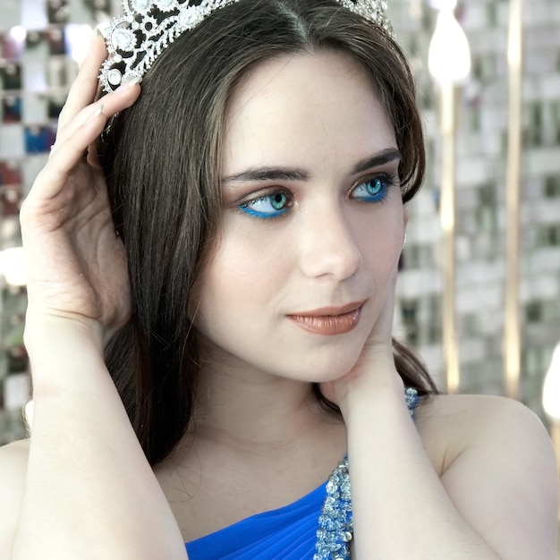 Młoda dziewczyna o niebieskich oczach w koronie i świątecznej niebieskiej sukience pozowanie na tle dekoracje noworoczne.