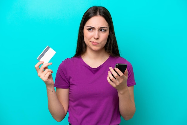 Młoda dziewczyna odizolowana na niebieskim tle, kupując telefonem komórkowym za pomocą karty kredytowej podczas myślenia