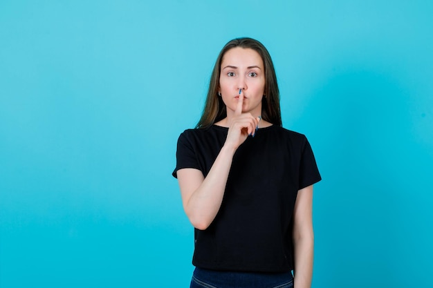 Młoda dziewczyna pokazuje gest ciszy, trzymając palec wskazujący na ustach na niebieskim tle