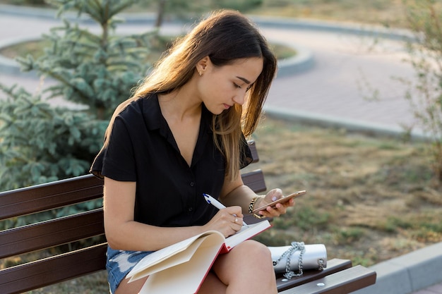 Młoda dziewczyna siedzi na ławce w parku i robi notatki Ubrana w stylu wolnym Biznes kobieta rozmawia przez telefon z klientami