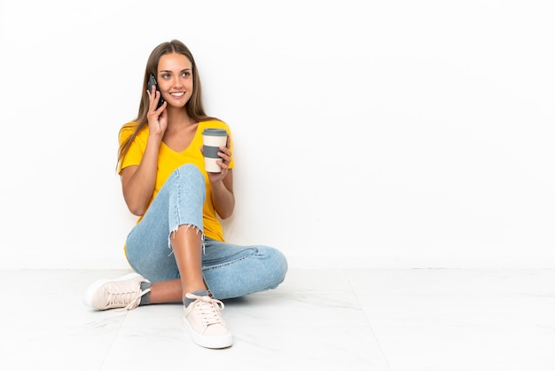 Młoda dziewczyna siedzi na podłodze trzymając kawę na wynos i telefon komórkowy