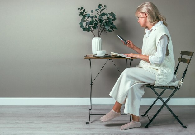 Młoda dziewczyna siedzi przy stoliku do kawy z telefonem w rękach, odwrócona od książki. Widok z boku.
