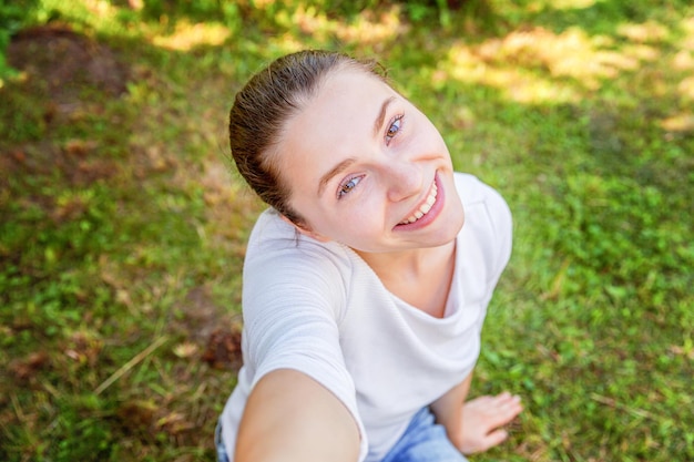 Młoda dziewczyna śmieszne wziąć selfie z rąk z telefonu, siedząc na tle zielonej trawie parku lub ogrodu. Portret młodej atrakcyjnej kobiety co selfie zdjęcie na smartfonie w letni dzień