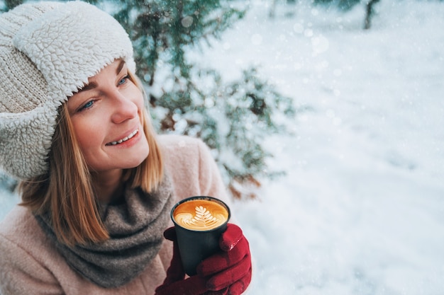 Zdjęcie młoda dziewczyna spaceruje i trzyma w rękach cappuccino w zaśnieżonym parku zimą