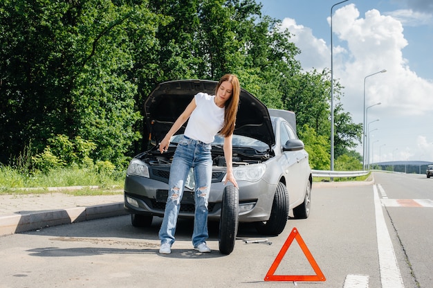 Zdjęcie młoda dziewczyna stoi przy zepsutym samochodzie na środku autostrady i próbuje zmienić złamane koło w upalny słoneczny dzień. awaria i awaria samochodu. czekam na pomoc.