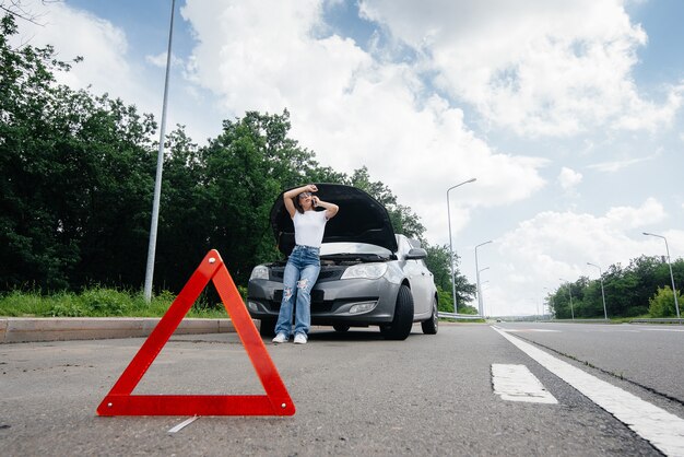 Młoda Dziewczyna Stoi Przy Zepsutym Samochodzie Na środku Autostrady I Woła O Pomoc Przez Telefon. Awaria I Awaria Samochodu. Czekam Na Pomoc.