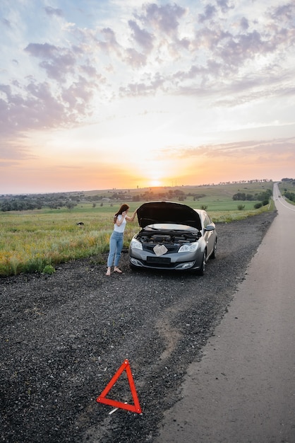 Młoda dziewczyna stoi w pobliżu zepsutego samochodu na środku autostrady o zachodzie słońca i próbuje wezwać pomoc przez telefon. Awaria i naprawa samochodu. Czekam na pomoc.