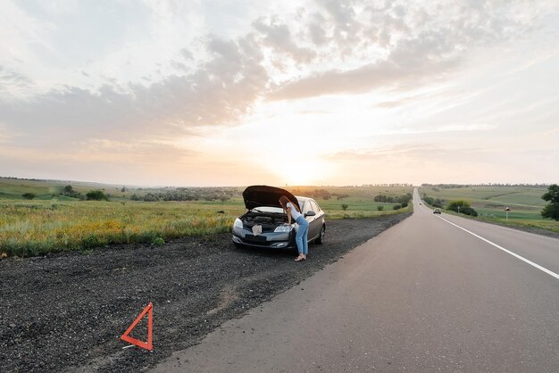 Młoda Dziewczyna Stoi W Pobliżu Zepsutego Samochodu Na środku Autostrady Podczas Zachodu Słońca I Próbuje Wezwać Pomoc Przez Telefon Czekając Na Pomoc Serwis Samochodowy Awaria Samochodu Na Drodze