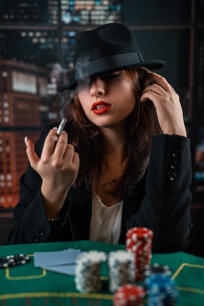 Młoda dziewczyna stylowo ubrana pali papierosa, wydmuchuje dym i gra w karty na stole na zielonym suknie pokera w kasynie