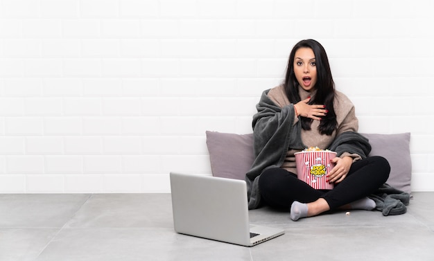 Młoda dziewczyna trzyma miskę popcorns i pokazuje film w laptopie, zaskoczony i zszokowany, patrząc w prawo