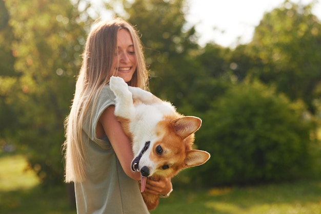 Młoda dziewczyna trzyma w ramionach wesołego i zabawnego walijskiego Corgi w parku w słoneczną pogodę koncepcja szczęśliwych psów