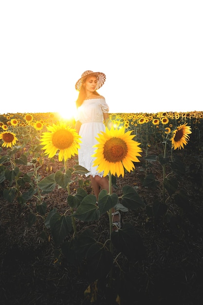 Młoda dziewczyna w białej sukni i kapeluszu na polu słoneczników o zachodzie słońca Portret kobiety o szczupłej sylwetce na tle żółtych kwiatów