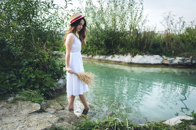 Młoda dziewczyna w białej sukni w słoneczny dzień wchodzi do jeziora. Dziewczyna w słomkowym kapeluszu w rzece o zachodzie słońca.