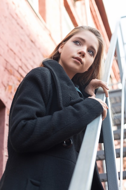 Młoda dziewczyna w czarnej płaszcz stojący na schodach z murem na tle. samotność.