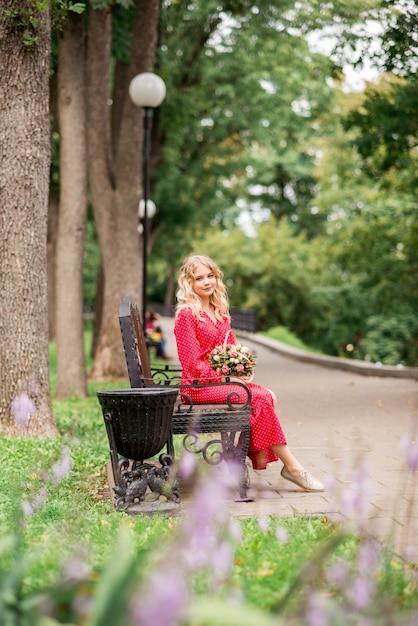 Młoda dziewczyna w czerwonej sukience siedzi na ławce w parku z bukietem