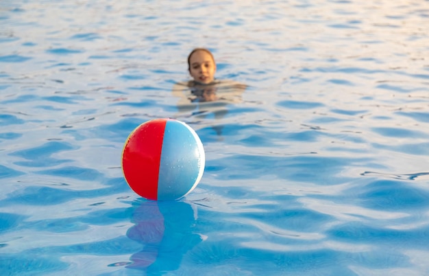 Młoda dziewczyna w jasnym stroju kąpielowym w panterkę pływa z jasnokolorową nadmuchiwaną piłką w ciemnoniebieskim basenie z czystą przezroczystą wodą w ciepły słoneczny letni wieczór