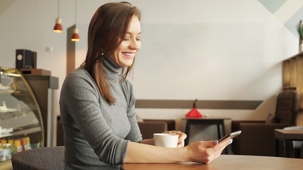 Młoda dziewczyna w kawiarni pisze wiadomość na telefonie i pije filiżankę kawy. Widok z boku, kopia miejsca