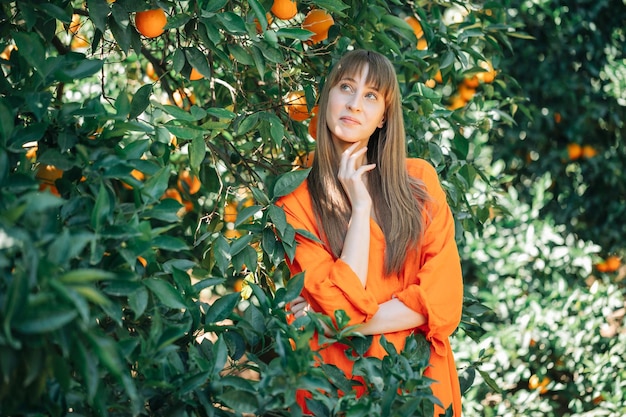 Młoda dziewczyna w pomarańczowej sukience patrzy w górę trzymając rękę pod brodą w pomarańczowym ogrodzie