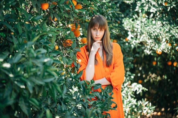 Młoda dziewczyna w pomarańczowej sukience patrzy w kamerę, trzymając rękę na brodzie w pomarańczowym ogrodzie