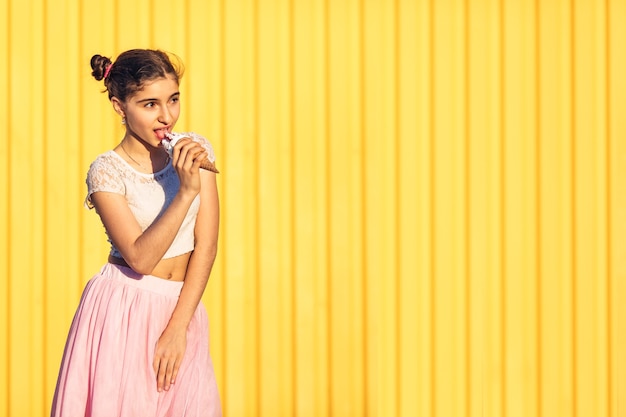 Młoda dziewczyna w różowej spódnicy je lody na żółtej ścianie