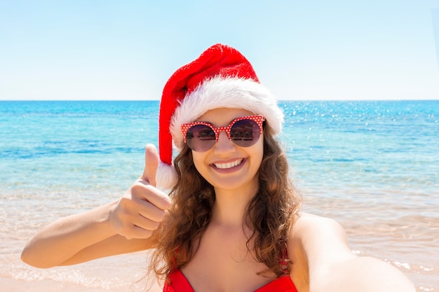 Zdjęcie młoda dziewczyna w santa hat pokazuje kciuk do góry i bawi się robienie zdjęć selfie smartfonem