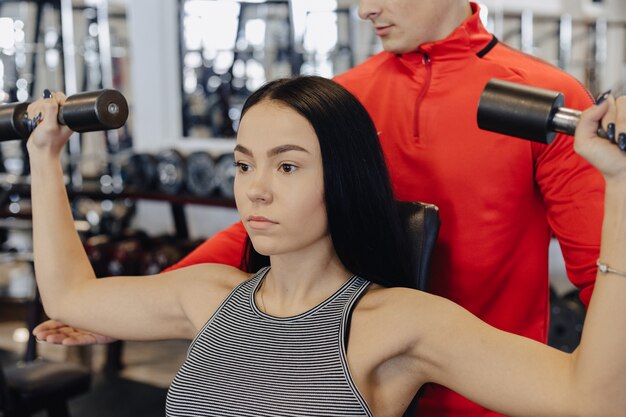 Młoda dziewczyna w sportowej odzieży gimnastycznej wykonuje ćwiczenia z hantlami, trener pomaga jej