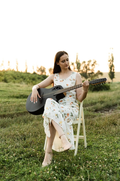 Młoda dziewczyna w sukience gra na gitarze, siedząc na wysokim krześle o zachodzie słońca. grać na gitarze. muzyk grający na instrumencie latem