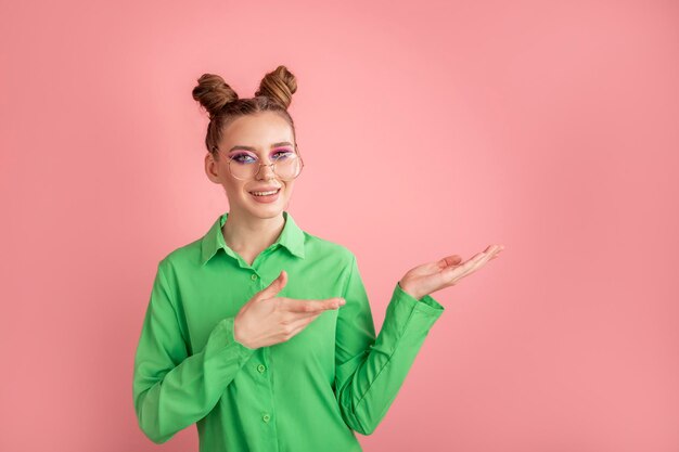 Młoda dziewczyna w zielonej koszuli i okularach na różowym tle pokazująca i wskazująca coś