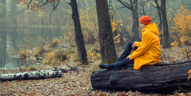 młoda dziewczyna w żółtym płaszczu przeciwdeszczowym siedzi na kłodzie na brzegu jeziora w lesie jesienią