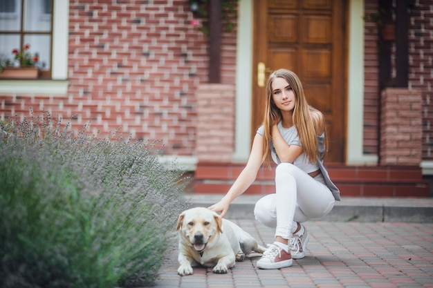 Zdjęcie młoda dziewczyna z aporterem na spacerze przed domem. atrakcyjna kobieta ubrana w stroje sportowe, głaszcząc labrador retriever i patrząc w kamerę.